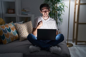 Canvas Print - Young hispanic man using laptop at home at night doing money gesture with hands, asking for salary payment, millionaire business