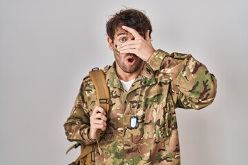 Wall Mural - Hispanic young man wearing camouflage army uniform peeking in shock covering face and eyes with hand, looking through fingers with embarrassed expression.