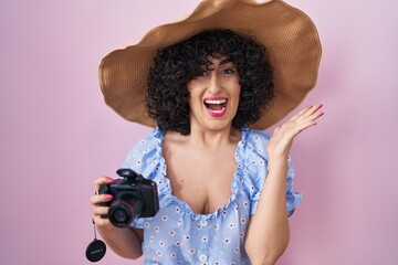 Wall Mural - Young brunette woman with curly hair using reflex camera celebrating victory with happy smile and winner expression with raised hands