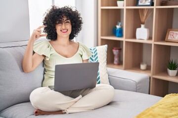 Canvas Print - Young brunette woman with curly hair using laptop sitting on the sofa at home smiling and confident gesturing with hand doing small size sign with fingers looking and the camera. measure concept.