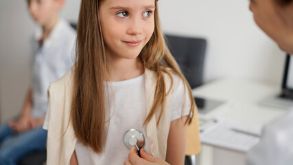 Wall Mural - Doctor and kid patient are in the clinic. Physician in white coat examining a smiling young girl with a stethoscope, close up. Medicine, therapy concept