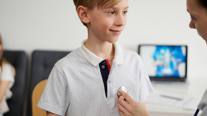 Wall Mural - Doctor and kid patient are in the clinic. Physician in white coat examining a smiling young boy with a stethoscope, close up. Medicine, therapy concept