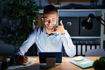 Poster - Young hispanic man working at the office at night smiling doing phone gesture with hand and fingers like talking on the telephone. communicating concepts.