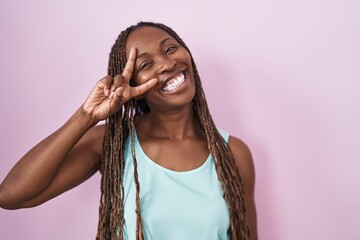 Poster - African american woman standing over pink background doing peace symbol with fingers over face, smiling cheerful showing victory