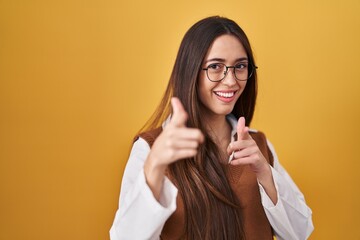 Sticker - Young brunette woman standing over yellow background wearing glasses pointing fingers to camera with happy and funny face. good energy and vibes.