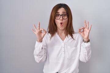 Wall Mural - Brunette woman standing over white isolated background looking surprised and shocked doing ok approval symbol with fingers. crazy expression