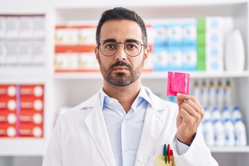 Wall Mural - Hispanic man with beard working at pharmacy drugstore holding condom thinking attitude and sober expression looking self confident