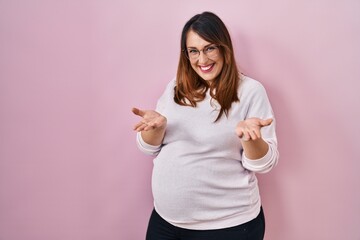 Poster - Pregnant woman standing over pink background smiling cheerful offering hands giving assistance and acceptance.