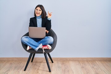 Poster - Young hispanic woman sitting on chair using computer laptop smiling with happy face winking at the camera doing victory sign with fingers. number two.