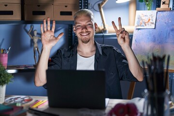 Sticker - Young caucasian man using laptop at night at art studio showing and pointing up with fingers number seven while smiling confident and happy.