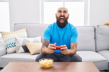 Canvas Print - Young hispanic man with beard and tattoos playing video game sitting on the sofa angry and mad screaming frustrated and furious, shouting with anger looking up.