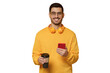 © Damir Khabirov - Young smiling man wearing yellow wireless headphones, holding takeaway coffee cup in one hand and phone in another
