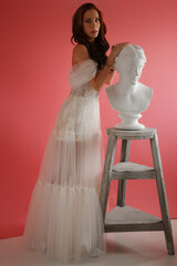 Beautiful brunette in a delicate white dress posing next to an ancient Greek bust on a pink background