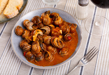 Roasted snails in the tomato sauce on a plate served with cutlery near to pieces of bread on the table in the Spanish cafeteria