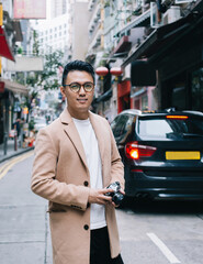 Smart ethnic well dressed guy standing at city street with photo camera