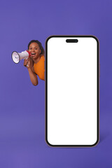 Wall Mural - Young admiring African American millennial woman with megaphone peeks out from behind giant phone and shouts loudly talking about advantages of new smartphone stands on purple background. White screen