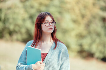 Wall Mural - Red-haired student girl in glasses with a notebook. Portrait outside.