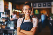 Portrait of a cheerful attractive satisfied smiling young woman with crossed arms and wearing apron working in a coffee shop