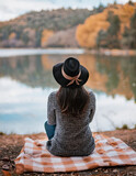 Fototapeta  - Giovane donna con cappello, all'aperto in riva al lago vista da dietro. Ambiente con colori autunnali