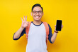 © Bangun Stock Photo - Smiling young Asian student man in casual clothes and glasses backpack showing smartphone blank screen and okay sign isolated on yellow background. Education in High School University College concept
