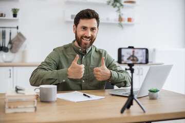 Wall Mural - Confident bearded man sitting at desk and working on wireless laptop while recording video on modern smartphone fixed on tripod showing thumbs up