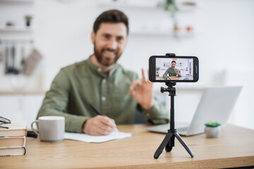 Wall Mural - Blurred background of smiling man taking notes while communicating with followers in social media during live streaming at home showing sign ok