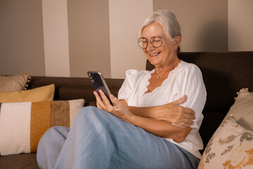 Wall Mural - Happy smiling senior white haired woman sitting on home sofa holding mobile phone. Elderly relaxed lady in video call