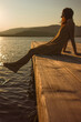 A contemplative young Caucasian woman sits alone on a lakeside pier, gazing at the water at sunset or dawn, in orange and vintage atmosphere.