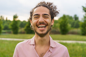 Outdoor portrait of a young man with curly hair tied back, laughing heartily with eyes squinted. He sports hoop earrings and a beard. The shot, taken in a park, has a shallow depth of field.