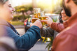 A diverse group of friends, including a gender fluid woman with vibrant short colored hair, raise their beer glasses in a toast at an outdoor sidewalk cafe, celebrating togetherness.