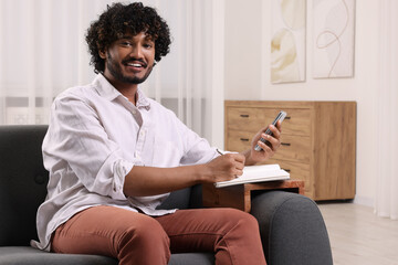 Sticker - Happy man using smartphone while writing in notebook on sofa armrest wooden table at home