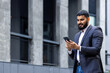 Young muslim man businessman standing on the street near an office building and using the phone, going to a meeting, calling, typing a message