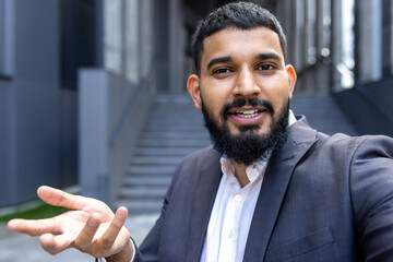 Wall Mural - Close-up portrait of a young Indian male businessman standing near an office building and talking on a video call from the phone