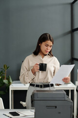 Fototapete - Young brunette woman in quiet luxury attire looking through financial document and having cup of coffee in office while standing by copier