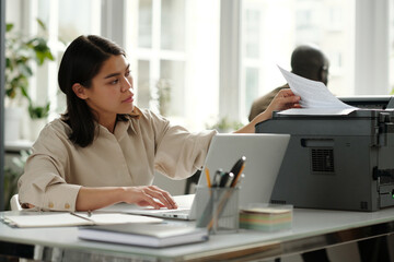 Fototapete - Busy young female accountant taking printed financial documents in xerox machine while sitting by workplace and typing on laptop keyboard