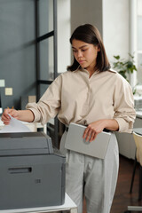 Fototapete - Young brunette businesswoman with tablet in hand standing in front of xerox machine and printing financial documents on working day