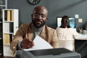 Fototapete - Focus on young African American employee taking paper from xerox tray while sitting in front of camera against female coworker with computer