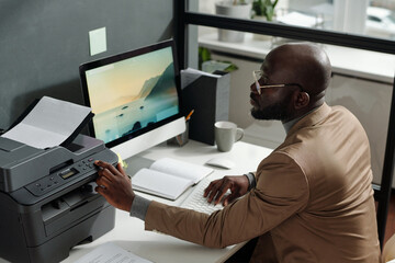 Fototapete - Young confident man in beige blazer pressing start button on panel of xerox machine while making some copies of financial documents