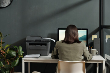 Fototapete - Rear view of young brunette secretary sitting on chair in front of computer screen in office and searching for online information in the internet