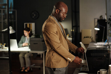 Fototapete - Side view of young African American businessman making cup of coffee in office while standing against female colleague with computer