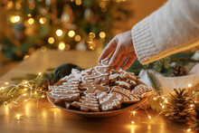 Merry Christmas! Hand Holding Gingerbread Cookie With Icing On Background Of Cookies In Plate On Table Against Christmas Tree Golden Lights. Atmospheric Christmas Holidays, Family Time