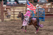 A cowgirl is riding a horse at a rodeo in a dirt arena. She is holding an American flag. The horse is brown and has sequence on its back . She has a pink blouse and a white hat. 