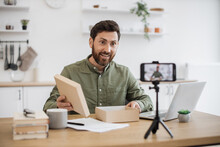 Caucasian young vlogger recording new video on smartphone about unboxing modern technologies. Bearded male person holding box and enjoying reviewing online purchase. Concept of working at home.