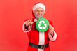 © khosrork - Satisfied positive elderly man with gray beard wearing santa claus costume pointing to camera with finger, showing green recycling sign. Indoor studio shot isolated on red background.