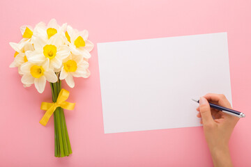 Wall Mural - Woman hand holding pen and writing on greeting card. Fresh white narcissus bouquet with yellow ribbon on pink table background. Pastel color. Empty place for text, quote or sayings. Top down view.