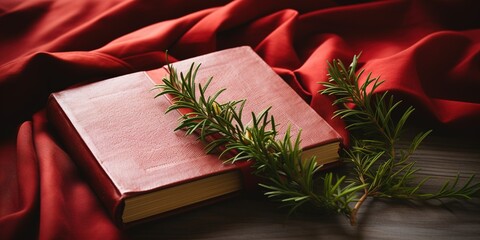 Wood cross laying on a closed red Christian bible with green fir branches on a white textile background. Christmas holidays.