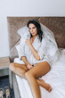 © shchus - A beautiful young brunette bride in lingerie and a shirt sits on the bed in the morning in a bedroom, hotel room. Wedding photography, portrait.