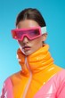 © Glittering Humanity - A striking woman sporting pink and yellow sunglasses exudes winter beauty as she poses in a ski mask and goggles, her vibrant orange jacket and spectacles adding to her fierce and stylish outdoor loo