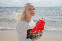 Romantic Beauty Portrait Of Young Happy Woman Enjoying Sea Outdoor. Beautiful Lady On Sea Beach Holding Little Red Sail Yacht