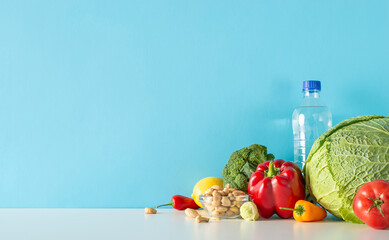 Wall Mural - Balanced living illustration: side view table composition comprising water bottle, cashew nuts, medley of vegetables, like broccoli, pepper and more set against blue wall for text or promo content